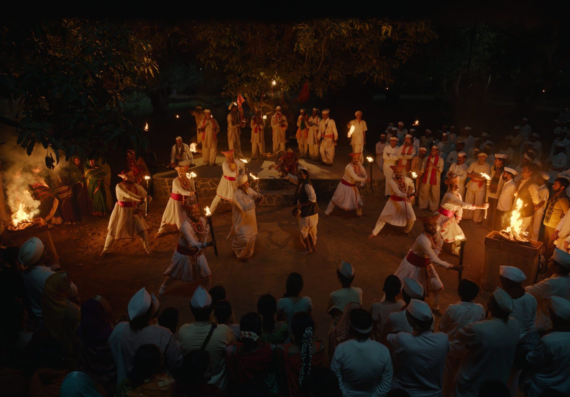 Overhead night view of Gondhali performers in white and red dancing with fire torches on a stone stage, surrounded by seated villagers.
