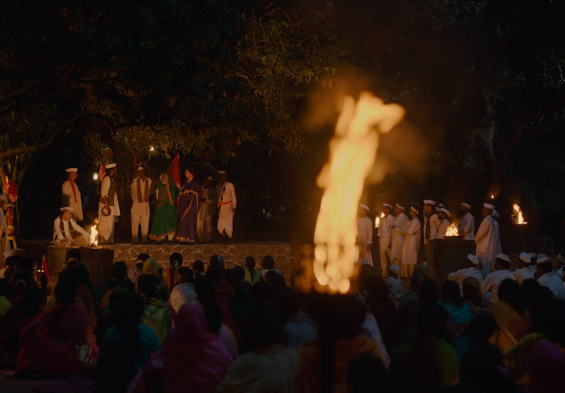 A large flame in the foreground, out of focus, with performers and red flags on a lit stone stage behind and a seated crowd in colourful saris.